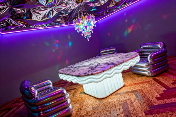 A contemporary dining room features a wavy-edged white marble table on a glowing, fluted acrylic base, surrounded by three metallic inflatable-style armchairs. Above, an iridescent crystal chandelier and a ceiling clad in crinkled mirrored mylar throw rainbow reflections across violet-lit walls, while perimeter LED strips add a neon wash and a warm mixed wood-and-cork herringbone floor grounds the vivid, psychedelic scene.