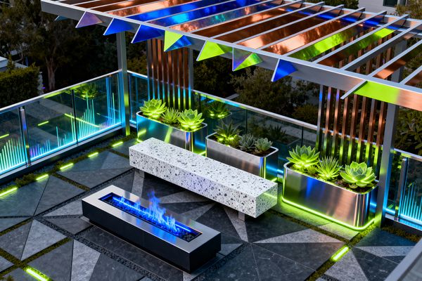 A rooftop-style patio with a linear blue flame fire pit, a long speckled white terrazzo bench, and stainless steel planters filled with succulents. A glass balustrade glows with cyan LEDs while green edge lighting traces the floor’s charcoal geometric pavers. Above, a pergola of brushed aluminum and copper fins with iridescent panels casts colorful reflections, creating a luminous cyberpunk atmosphere.
