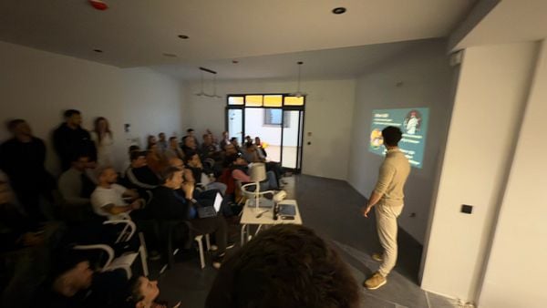 A tech meetup is taking place in a modern, well-lit room with a speaker presenting to an engaged audience.