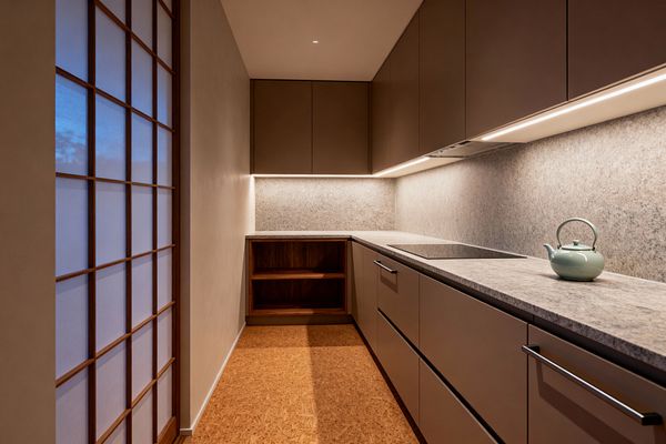 A narrow, minimalist galley kitchen with matte taupe slab cabinets and stainless bar pulls, featuring a light gray stone countertop and full-height stone backsplash illuminated by continuous under-cabinet LEDs. A flush black induction cooktop, an integrated hood, open walnut shelving, and cork flooring keep the space simple, while a wood-framed shoji screen along one wall softens the room; a pale green kettle sits on the counter.