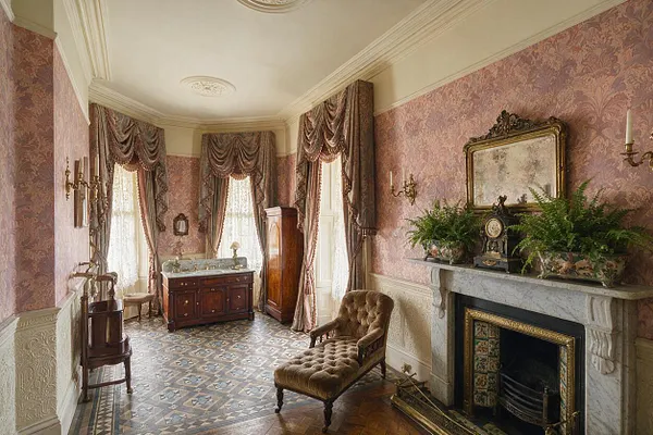A Victorian-style master bathroom with pink patterned wallpaper, tall draped windows, a carved marble fireplace, and ornate brass sconces. The room includes a dark wood vanity with a marble top, a matching cabinet, a tufted chaise lounge, and rich patterned tile flooring.