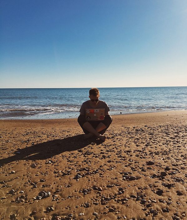 A person is working on a laptop while sitting on a beach.