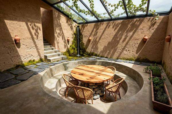 A sunken circular concrete dining pit features a round solid-wood table set on a base of stacked river stones, surrounded by four rattan chairs. The space has rough, sand-colored earthen walls with terracotta wall pots and a long planter of herbs. Overhead, a glass roof with trailing vines casts leaf shadows, and a narrow waterfall descends into moss near a slate path. Concrete steps lead up to a doorway, creating a serene, nature-focused dining area.
