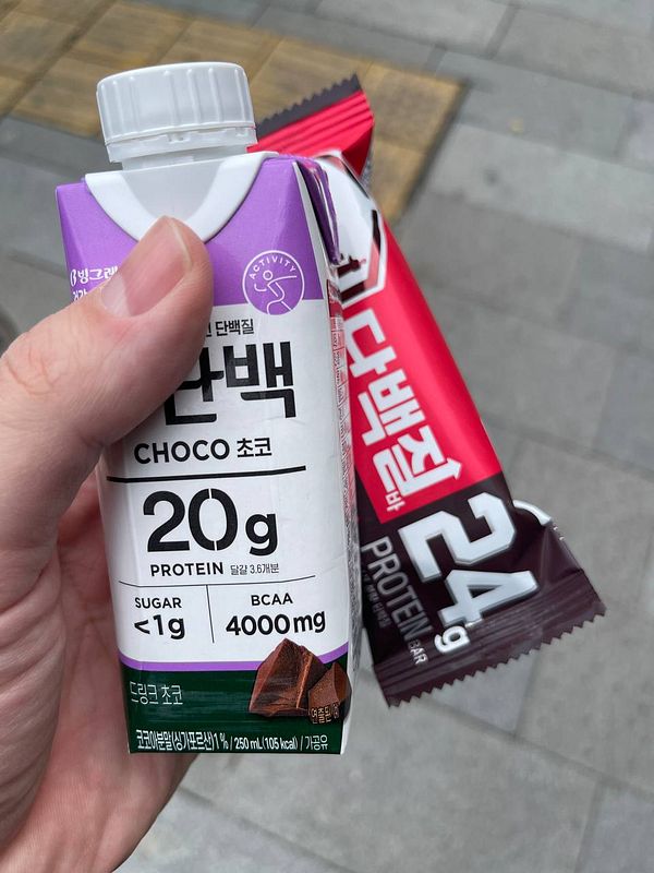 A hand holds a protein drink and a protein bar against a blurred outdoor background.