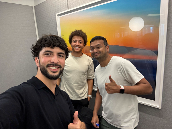 Three individuals pose together in an office setting, smiling and giving thumbs up.