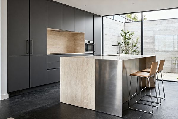 A minimalist kitchen features matte charcoal flat-front cabinetry with discreet stainless handles, a built-in stainless wall oven, and a warm travertine stone backsplash and countertop niche. A sculptural island pairs a travertine waterfall end with a brushed stainless steel counter and integrated sink, accompanied by two tan leather bar stools; dark large-format stone flooring and full-height black-framed windows bring generous natural light and courtyard views.