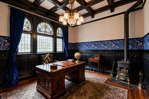 Victorian-style home office with a carved mahogany desk on a patterned rug, accessorized by a leather chair, brass telescope, and globe. A dark coffered ceiling and black paneled wainscoting frame arched stained-glass windows with royal blue velvet curtains; a brass-and-glass chandelier hangs above, and a cast-iron stove with gold accents sits on a hearth to the right beside a narrow side table.