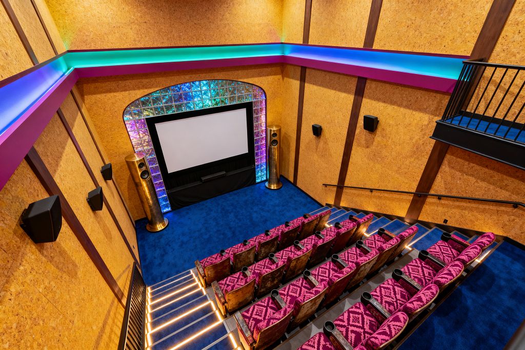 A home theater with steep, tiered rows of fuchsia diamond-patterned seats faces a large screen. The screen is framed by an iridescent glass-block arch with tall gold speakers on each side, set over a cobalt blue carpet; cork-paneled walls with dark wood battens, LED-lit stair risers, magenta trim with teal-to-blue cove lighting, multiple black wall speakers, and a black metal balcony rail complete the bold maximalist space.