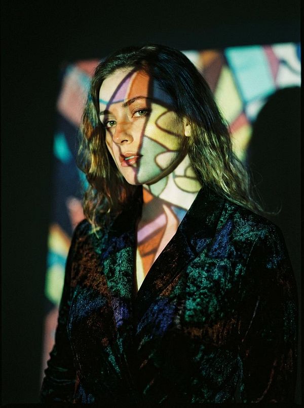 A woman stands against a dark background, illuminated by colorful projections that create intricate patterns on her face and clothing.