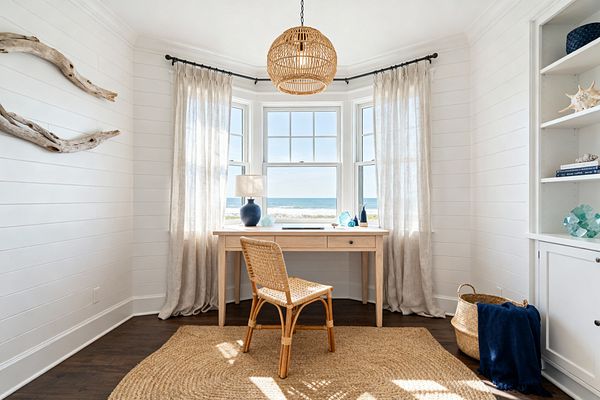 A bright coastal home office centers on a light wood desk set in a bay window overlooking the ocean. White shiplap walls, sheer beige linen curtains on a black rod, and a woven rattan pendant create an airy feel. A rattan chair sits on a round jute rug atop dark hardwood floors. Driftwood art hangs on the left wall, while built-in white shelves display sea-glass decor and shells. A blue ceramic lamp and a seagrass basket with a navy throw add color.