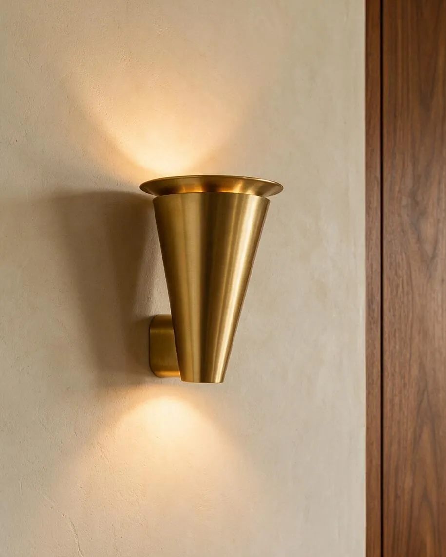 A brass wall sconce with a tapered conical shape casts warm light upward and downward on a textured beige wall. A vertical wood panel sits to the right, adding contrast and a mid-century modern feel.
