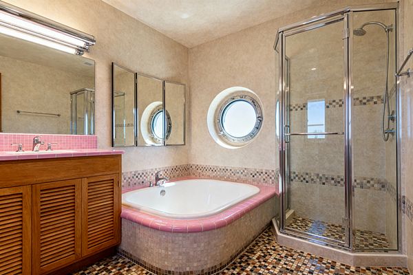 A mid-century bathroom features a white soaking tub wrapped with pink ceramic bullnose tile and a mosaic-clad apron beside a chrome-framed glass shower lined in beige tile with a small mosaic border. A warm wood louvered vanity with a pink mosaic countertop sits under tri-panel mirrored cabinets and a chrome fluorescent light, while a round porthole window above the tub brings daylight to a floor of multitone beige and brown mosaic tile.