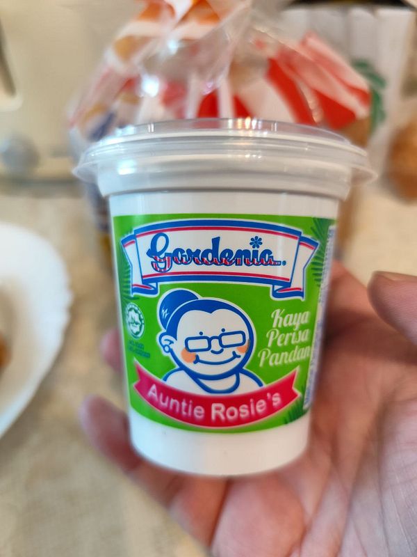 A container of kaya spread is held in a hand, featuring a colorful label.