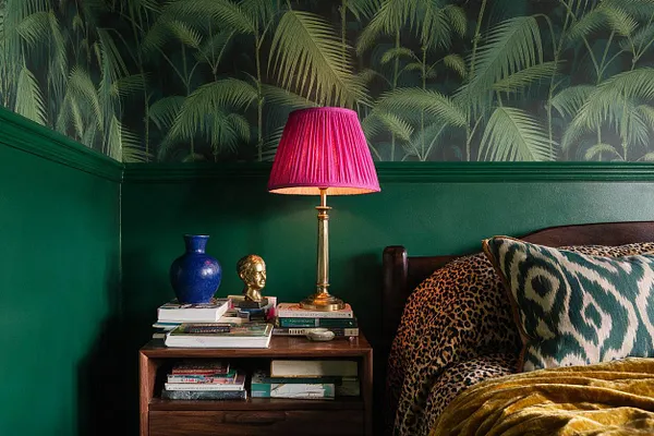 A maximalist bedroom vignette with deep green walls and tropical palm wallpaper above dark molding. A wooden bedside table holds stacked books, a cobalt vase, and a small gold bust under a brass lamp with a bright magenta shade, beside a dark wood bed layered with leopard and ikat-style patterned bedding.