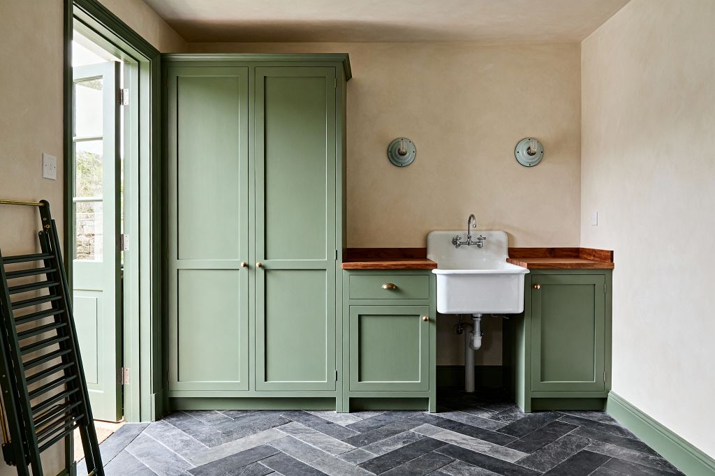 A light-filled farmhouse laundry room with sage green Shaker-style cabinets and matching door and trim. A tall cupboard sits beside base units topped with a warm wood butcher-block counter. At the center is a deep white utility sink with an integrated backsplash and a wall-mounted chrome faucet, with exposed plumbing below. The floor is charcoal and gray slate laid in a herringbone pattern. Creamy plaster walls, two simple green sconces, and brass hardware complete the space.