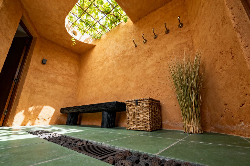 An earthy mudroom with rough terracotta plaster walls features a black timber bench, six brass wall hooks, a woven rattan storage basket, and a bundle of tall reeds. Green ceramic floor tiles flank a central drain filled with porous lava stones, while a round skylight with a vine-covered metal trellis pours dappled natural light across the space.