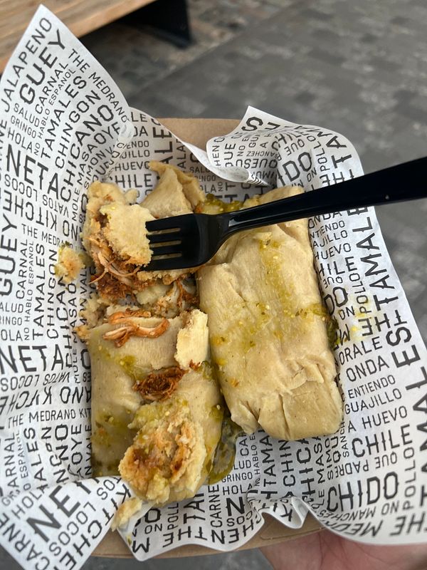 A hand holds a tray containing two tamales topped with green sauce.
