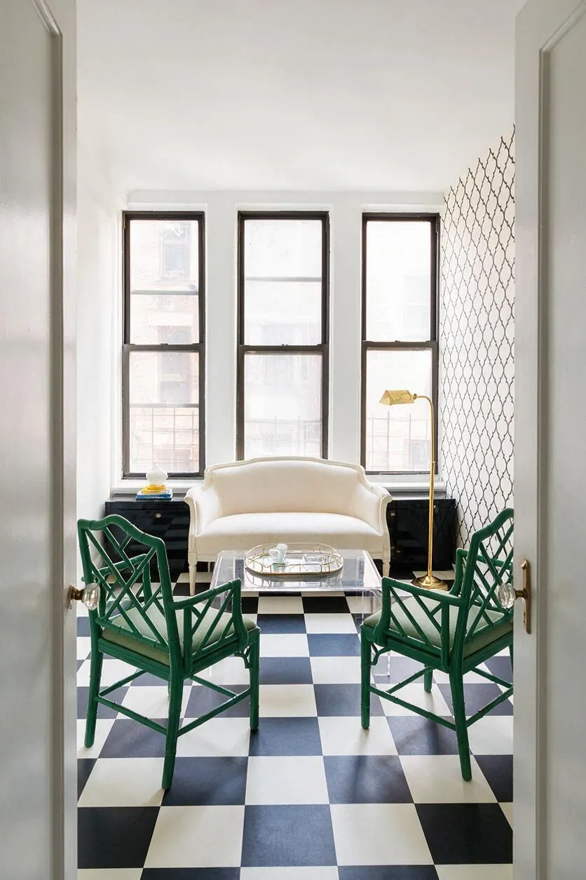 A bright sunroom styled in Hollywood Regency fashion, featuring a black-and-white checkerboard floor, three tall windows, a cream loveseat, two emerald green bamboo-style chairs, a glass coffee table, and a brass floor lamp. Patterned wallpaper on one side and glossy black window bases add contrast.