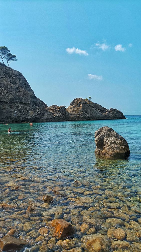 A serene coastal scene featuring clear water, rocky formations, and a couple of swimmers.