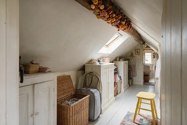 A narrow attic closet with sloped white ceilings, exposed wood beam, and soft daylight from a skylight. White painted cabinets line the walls, paired with wicker baskets, a folded textile bin, hanging dresses, a yellow stool, and dried floral garlands overhead, creating a cozy cottagecore look.