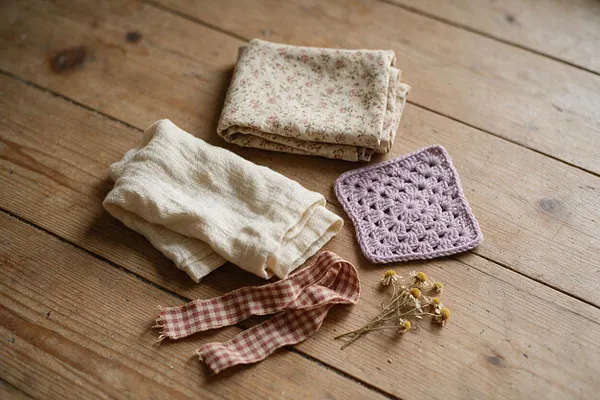 A flat lay of cottagecore materials arranged on a rustic wooden surface, including folded floral cotton fabric, cream linen, a lavender crocheted square, a red gingham ribbon, and a small sprig of dried flowers. The warm wood grain and muted colors create a cozy handmade feel.
