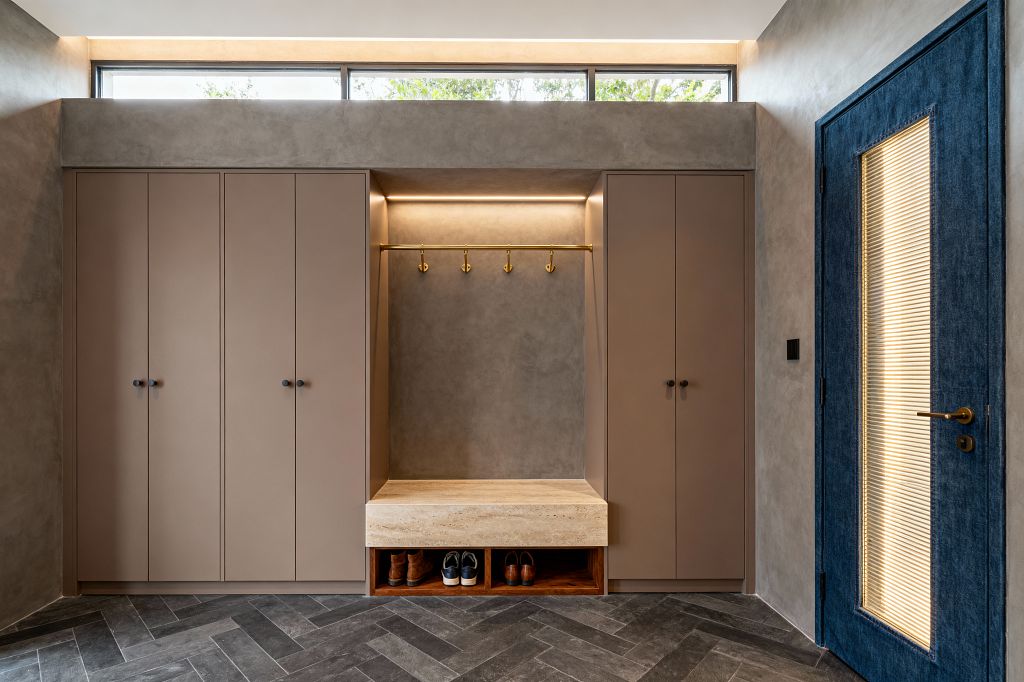 Contemporary mudroom with symmetrical taupe cabinets flanking a central niche. A thick travertine bench sits above walnut cubbies for shoes, with a brass rod and five hooks beneath a concealed LED strip. Walls are warm gray microcement and the floor is charcoal herringbone tile. A long clerestory window and cove lighting add daylight, while a deep blue door with ribbed glass and a brass lever punctuates the right side.