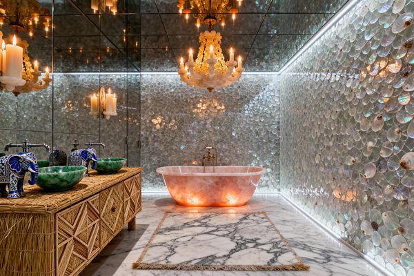 An opulent bathroom features mirrored walls and ceiling reflecting candle-style chandeliers over iridescent shell-tile walls. A freestanding pink onyx bathtub glows from below on a white marble floor with a fringed rug, while a woven rattan vanity holds two green stone vessel sinks with chrome taps and blue-and-white elephant accents; a brass tub filler stands beside the bath, and linear LEDs trace the room’s perimeter.