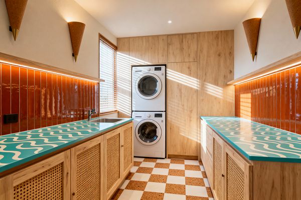A galley-style laundry room features aqua countertops with white boomerang waves above cane-front light oak cabinets, flanking a central aisle. Glossy terracotta vertical tiles run the length of both walls under a slim LED strip. At the end, a stacked white washer and dryer are framed by full-height oak paneling beside a window with wood blinds. The floor is a cork-and-white checkerboard, and cone sconces with brass tips cast warm light over a stainless sink and chrome faucet.