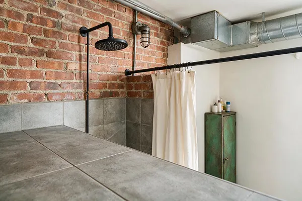 A master bathroom with an industrial style featuring an exposed red brick wall, gray tile surround, black metal shower fixtures, and visible galvanized ductwork near the ceiling. A cream shower curtain hangs beside a weathered green metal cabinet, creating a raw yet refined loft-like atmosphere.