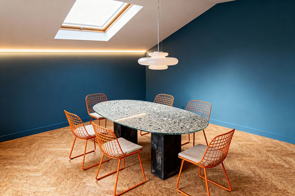 A modern dining room features deep teal walls and a sloped ceiling with a skylight and a slim LED strip. At center sits an oval terrazzo table on two blackened metal pedestals, encircled by six orange powder-coated wire chairs with light beige cushions, set on a warm herringbone cork floor, and lit by a white opal-glass pendant.