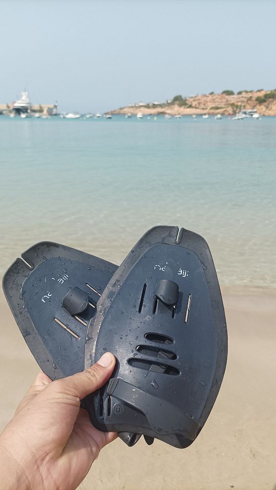 A person holds a pair of swim fins against a scenic beach backdrop.