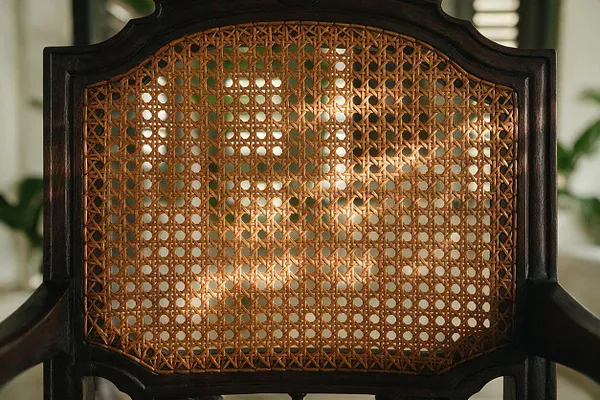 A close-up of a tropical-style chair showing a dark carved wood frame and a tightly woven cane backrest in warm honey tones. Soft natural light highlights the intricate lattice pattern, with blurred greenery in the background suggesting an airy indoor setting.