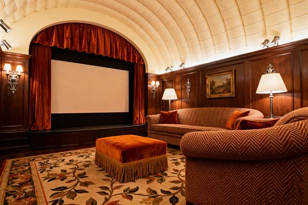 A traditional home theater shows a large projection screen framed by arched burnt-orange velvet curtains beneath a cream barrel-vaulted ceiling. Dark walnut wall paneling surrounds herringbone-upholstered sofas with rust velvet pillows, a fringed velvet ottoman, and a floral wool rug. Nickel sconces, pleated-shade lamps, and subtle uplights warm the woodwork and ceiling, creating an intimate, classic cinema atmosphere.