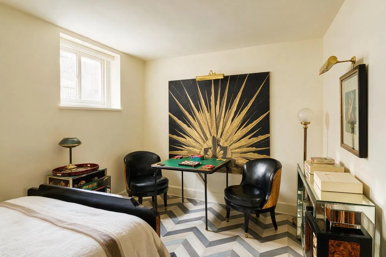 A compact Art Deco-inspired game room with a small card table at the center, surrounded by two black leather chairs. A large black artwork with a gold sunburst dominates the back wall, while mirrored side tables, brass lamps, and a bold chevron-patterned floor add glamour and geometric rhythm.