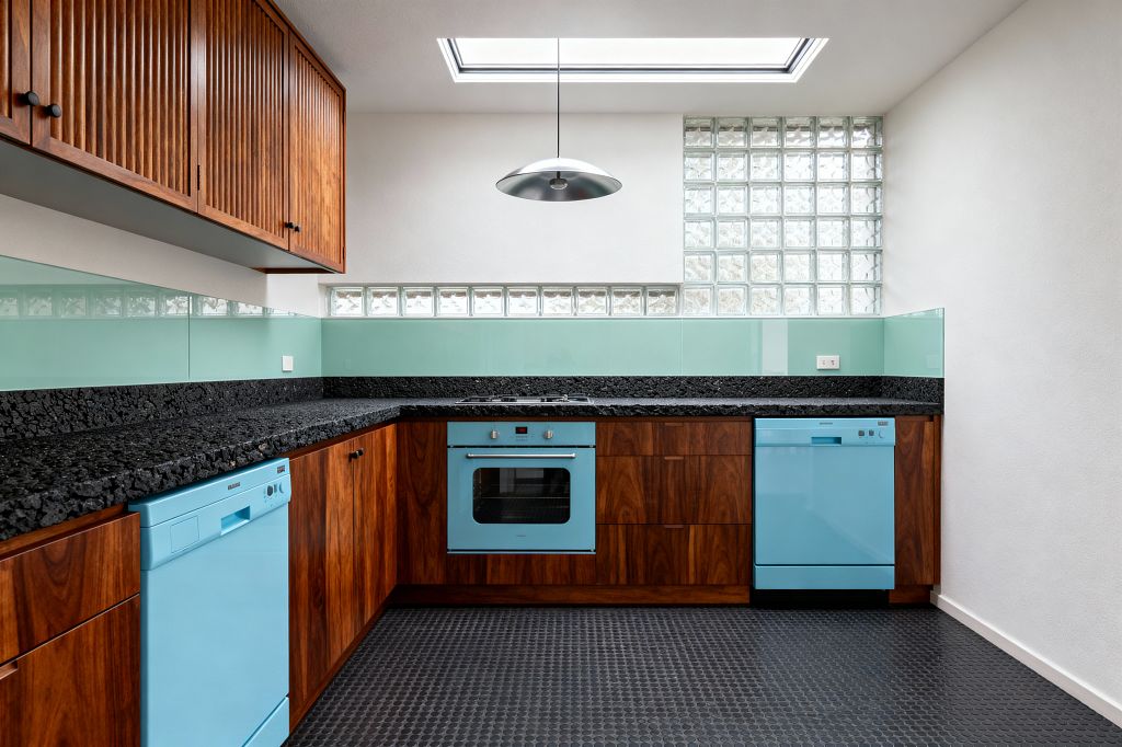 A mid-century inspired utility room with L-shaped walnut cabinetry and fluted upper doors beneath a large skylight. Retro aqua-front base appliances and a matching built-in oven sit below a thick, textured black stone countertop, with a mint back-painted glass backsplash and glass-block windows diffusing light. The floor is black penny-round tile, and a brushed aluminum saucer pendant hangs centrally for task lighting.