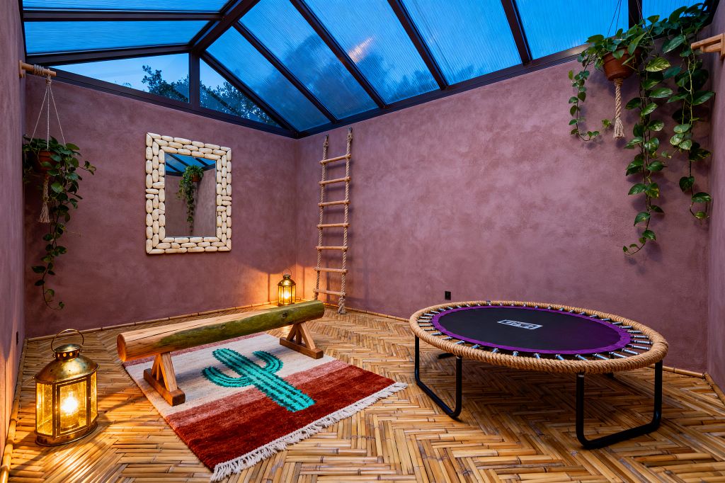 A bohemian home gym with mauve stucco walls sits under a sloped, blue-tinted skylight. A rope-edged mini trampoline, a natural log balance beam on a cactus-patterned rug, brass lanterns casting warm light, a pebble-framed wall mirror, a rope ladder in the corner, bamboo herringbone flooring, and trailing plants in terracotta hanging pots create an earthy, eclectic workout space.