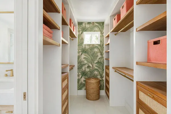 A narrow walk-in closet with white built-in shelving and warm wood shelves on both sides. Coral storage bins and woven drawers add color and texture, while a palm leaf wallpaper accent wall and a small window create a bright tropical feel.