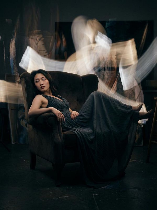 A woman in a flowing dark dress lounges on a plush armchair, surrounded by ethereal light trails in a dimly lit studio.