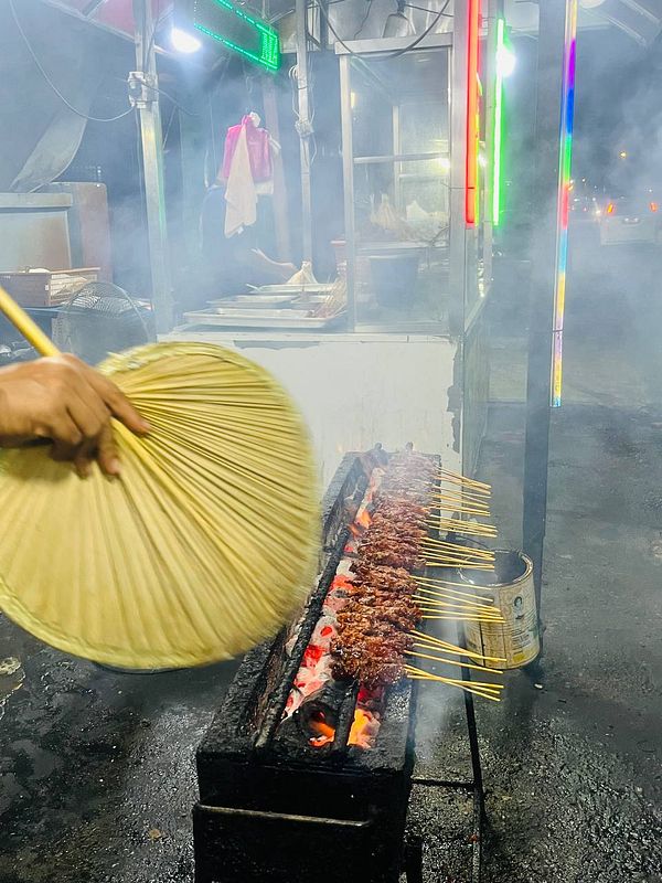 A street vendor is grilling satay skewers over an open flame while fanning the smoke away.