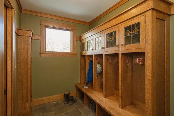 A Craftsman-style entryway with built-in oak storage cubbies and a bench lining one wall. The upper cabinet doors feature small leaded glass panels, while the lower open cubbies hold hooks for coats and bags; olive green walls and natural wood trim create a warm, traditional feel.