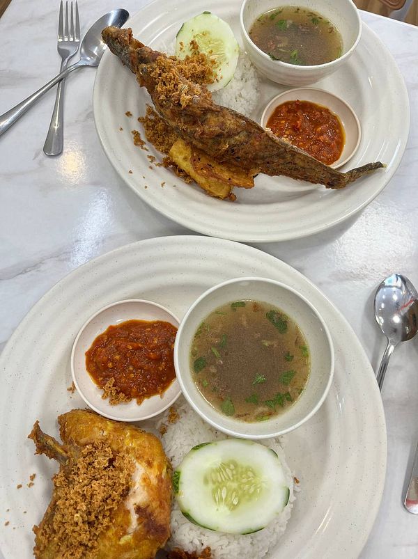 Two plates of Nasi Ayam Penyet featuring fried chicken and fish, served with rice, cucumber, sambal, and soup.