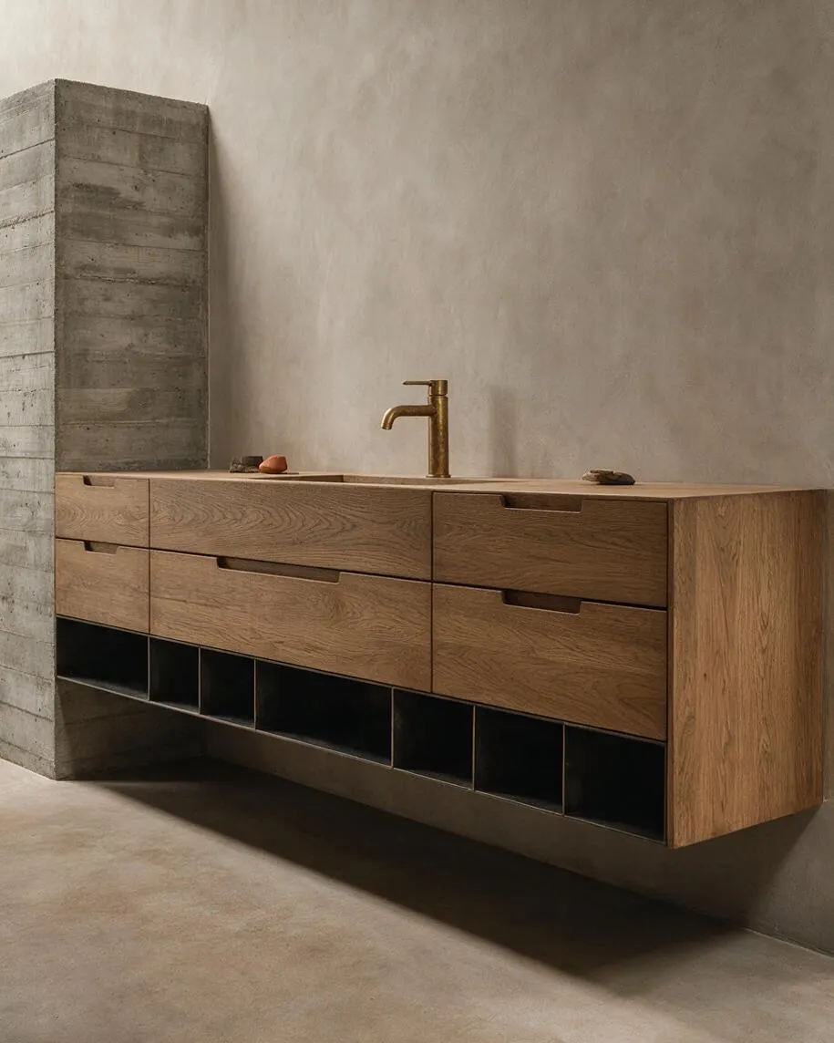 A master bathroom with a floating wooden vanity featuring multiple drawers and open cubbies below, paired with a brass faucet and set against a smooth beige wall. A raw concrete partition stands to the left, reinforcing the minimalist Brutalist look.