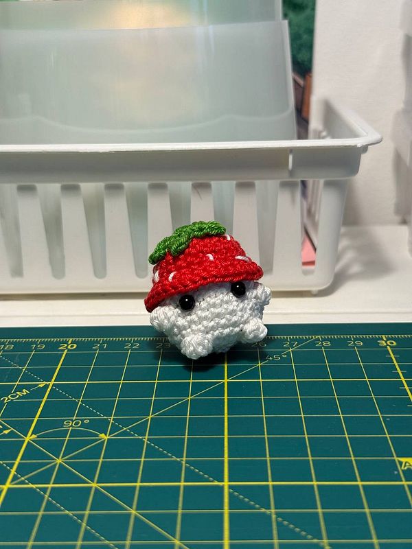 A crocheted critter resembling a strawberry sits on a green cutting mat.