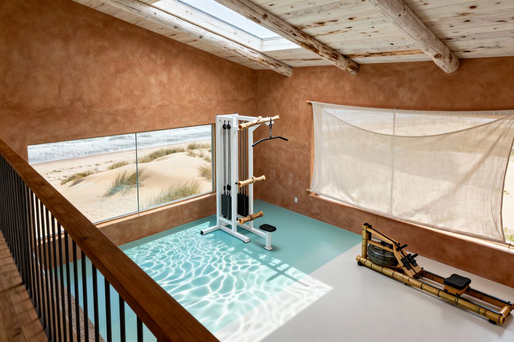 A coastal home gym overlooks sand dunes and the ocean through a wide picture window, with terracotta limewashed walls and a whitewashed timber ceiling with exposed log beams and a skylight. The sea-glass epoxy floor catches rippled sunlight, framing a white cable machine with driftwood-style handles and a wooden water rower; a sheer linen shade filters light, and a slim black railing borders the mezzanine.