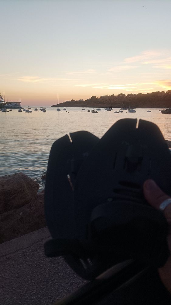 A pair of swim fins is held in front of a serene waterfront scene at sunset.