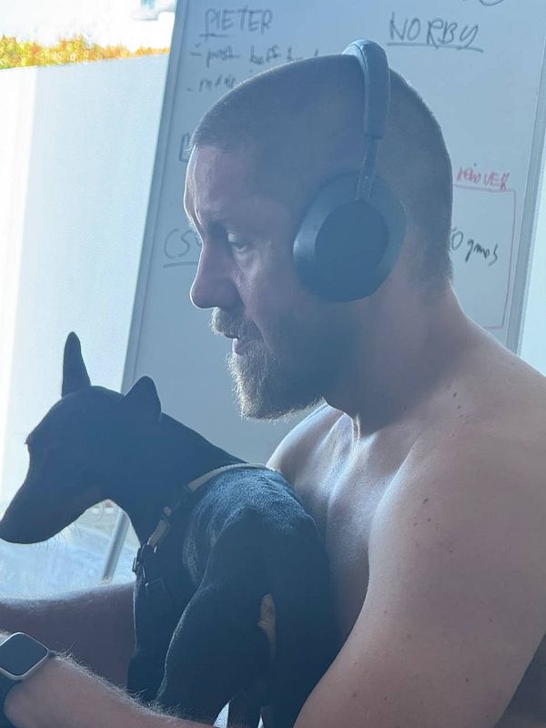A shirtless man wearing headphones sits with a small black dog on his lap.