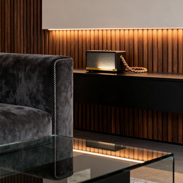 Close-up of a contemporary media room featuring vertical walnut slat wall paneling with warm LED backlighting, a floating matte-black media console, and a compact speaker with brass trim and a braided rope handle. In the foreground, a charcoal velvet sofa with piped edges sits beside a clear glass coffee table over polished concrete flooring, creating a moody, refined atmosphere.