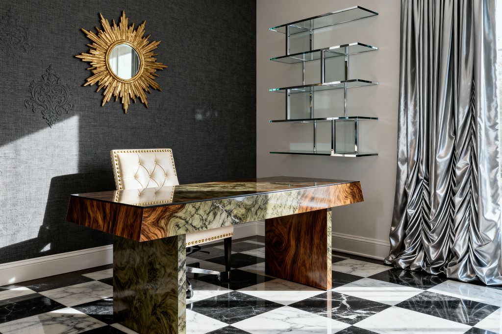 A glamorous home office features a glossy desk combining green-veined marble and rich walnut, centered on a black-and-white checkerboard marble floor. A tufted cream leather chair with brass nailheads faces a charcoal textured wall with subtle damask embossing and a gold sunburst mirror. On the right, chrome-framed glass shelves float over a soft gray wall beside voluminous silver satin drapes, with natural light highlighting the reflective finishes.