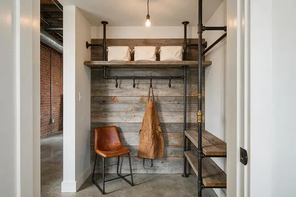 A narrow industrial-style entryway with a rustic wood slat wall, black metal pipe shelving and coat hooks, and a single exposed bulb overhead. A tan apron hangs on the hooks, a brown leather chair sits to the left, and built-in wood shelves line the right side, with white walls framing the space.