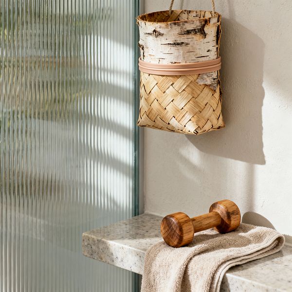 A Scandinavian-style home gym vignette shows a light gray terrazzo floating shelf against warm off‑white plaster, illuminated by sunlight filtered through a vertical reeded glass screen. A honey‑toned wooden dumbbell rests on a sand‑beige cotton towel, while a small hanging basket woven from natural straw with birch‑bark detail provides storage, all in a calm, neutral palette.